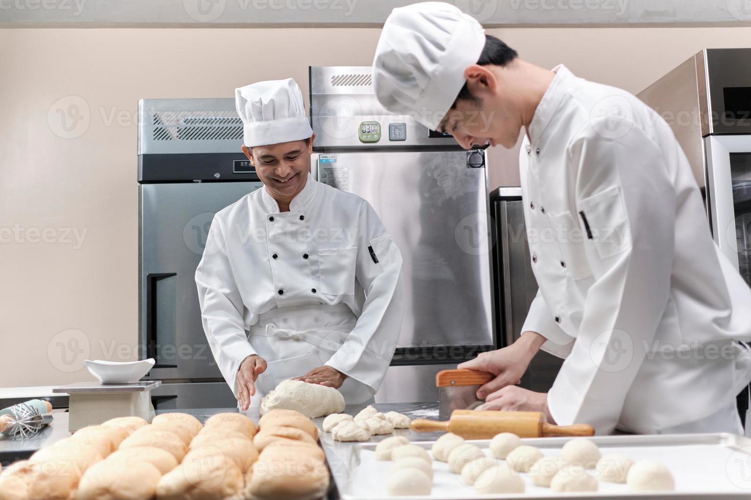 dos chefs asiáticos profesionales con uniformes y delantales de cocinero blanco están amasando masa de pastelería y huevos, preparando pan y comida fresca de panadería, horneando en el horno en la cocina de acero inoxidable del restaurante. foto