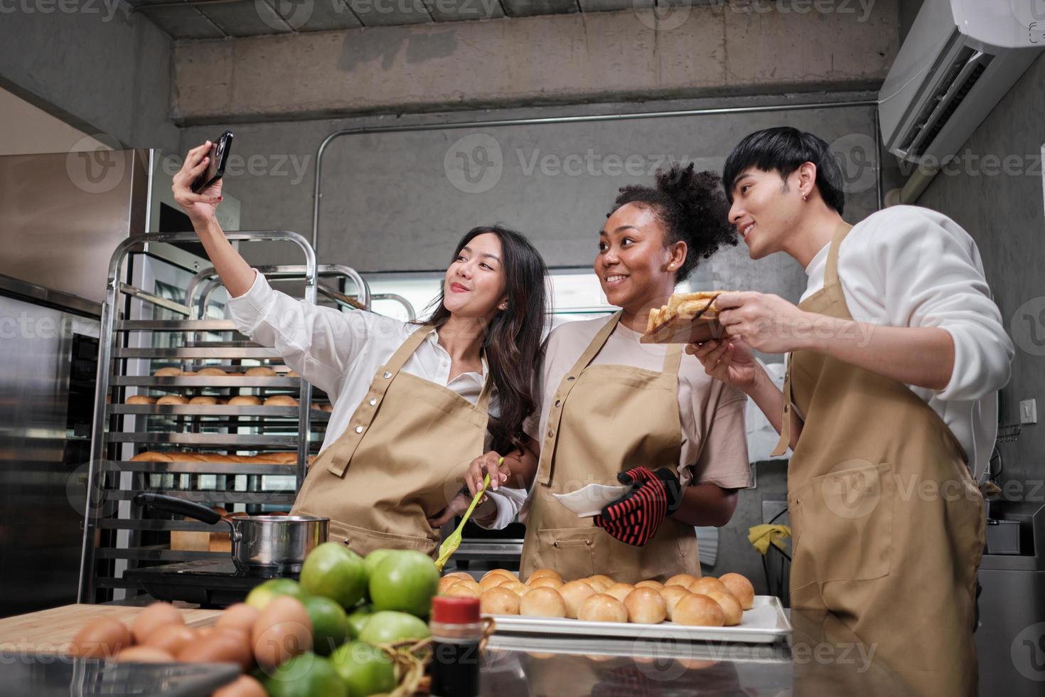 tres jóvenes estudiantes en la clase de cocina usan delantales para disfrutar mientras toman una foto selfie con el teléfono móvil en la cocina, sonríen y ríen, preparan huevos y frutas, aprenden juntos un curso culinario divertido.