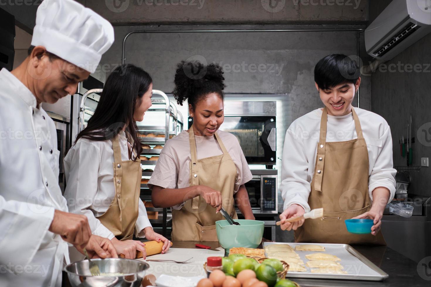 curso de cocina, chef masculino senior en uniforme enseña a jóvenes estudiantes de cocina, cepilla la masa de pastelería con crema de huevos, prepara ingredientes para alimentos de panadería, tartas de frutas en la cocina de acero inoxidable. foto