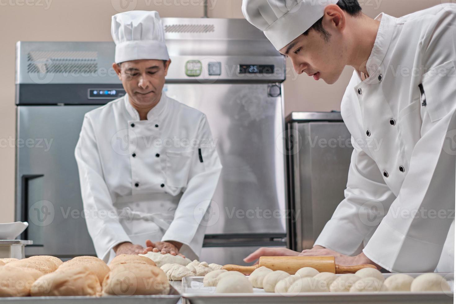 Two professional Asian male chefs in white cook uniforms and aprons are kneading pastry dough and eggs, preparing bread and fresh bakery food, baking in oven at stainless steel kitchen of restaurant. photo