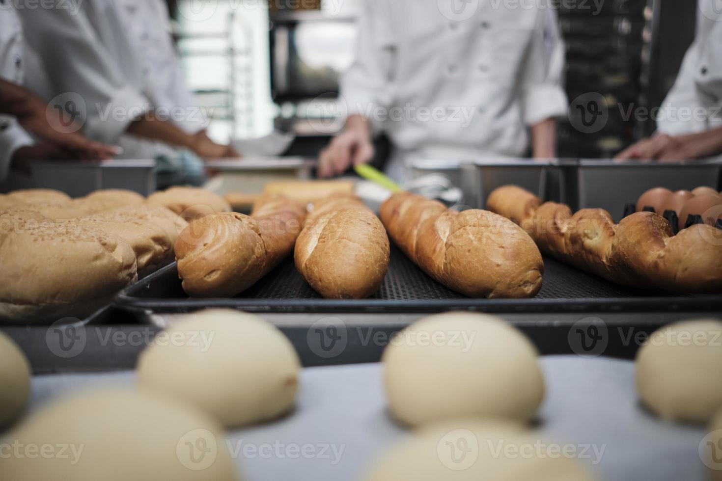 toma de primer plano y enfoque selectivo en baguettes y pan delicioso frente al equipo de chefs de uniforme blanco, amasando masa de pastelería cruda, preparando comida fresca de panadería, horneando en el horno en la cocina del restaurante. foto