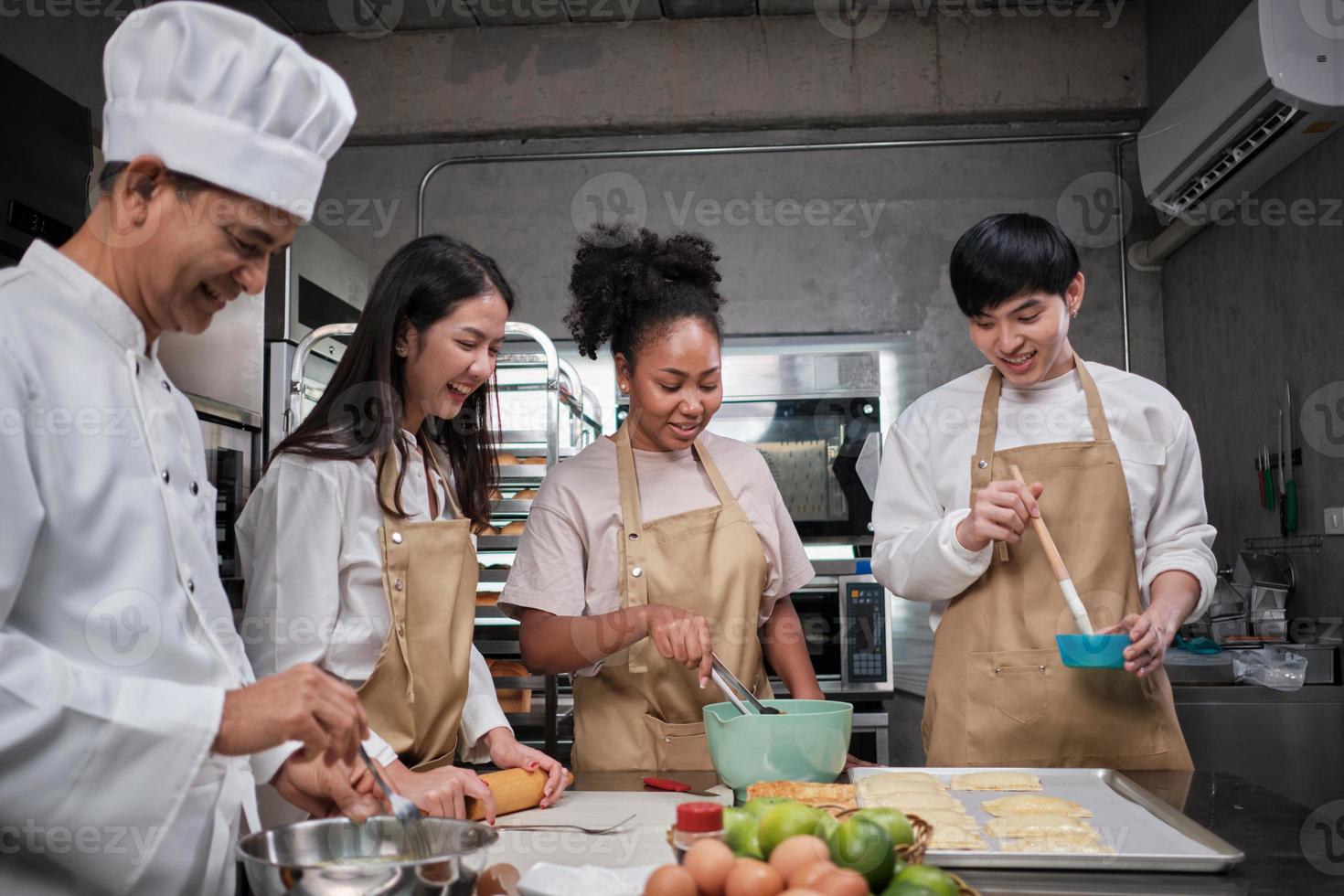 curso de cocina, chef masculino senior en uniforme enseña a jóvenes estudiantes de cocina, cepilla la masa de pastelería con crema de huevos, prepara ingredientes para alimentos de panadería, tartas de frutas en la cocina de acero inoxidable. foto