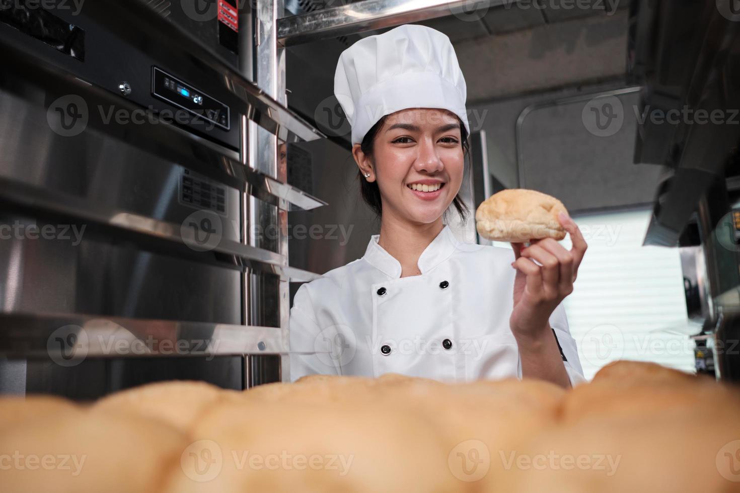 Young Asian female chef in white cook uniform and hat showing tray of fresh tasty bread with smile, looking at camera, happy with his baked food products, professional job at stainless steel kitchen. photo