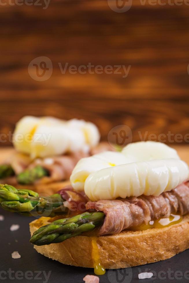 tostadas a la parrilla con espárragos en tocino y huevo escalfado sobre fondo de madera foto