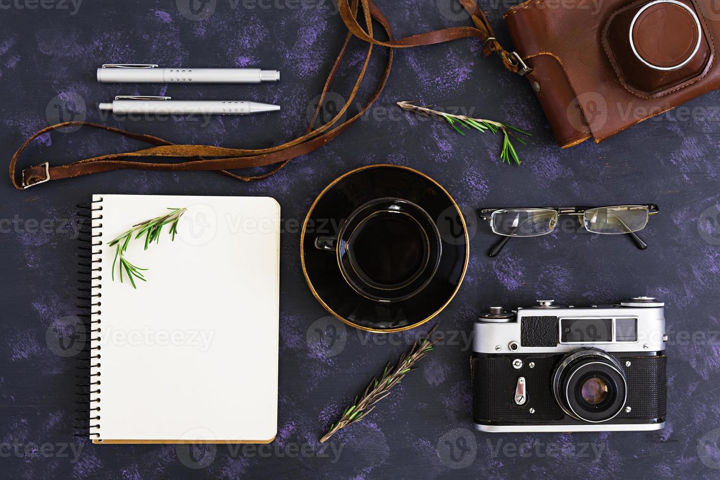 Flat lay, top view office table desk. Desk workspace with retro camera, diary, pen, glasses, case, cup of coffee, rosemary on dark background. photo