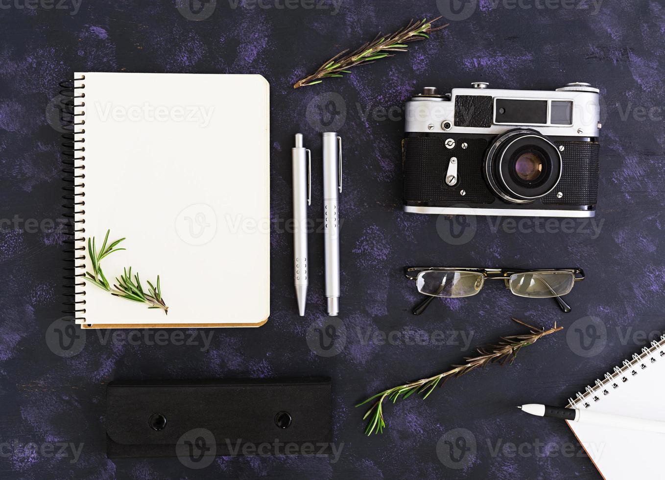 Flat lay, top view office table desk. Desk workspace with retro camera, diary, pen, glasses, case, rosemary on dark background. photo