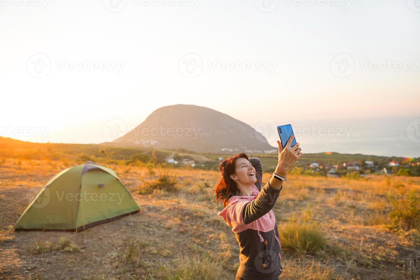 Woman takes photos of the sunrise in the mountains on her phone. Selfie in rising sun. Panoramic view on sea and Ayu-Dag. Camping, outdoor activities, sports mountain hiking, family travel. Crimea.