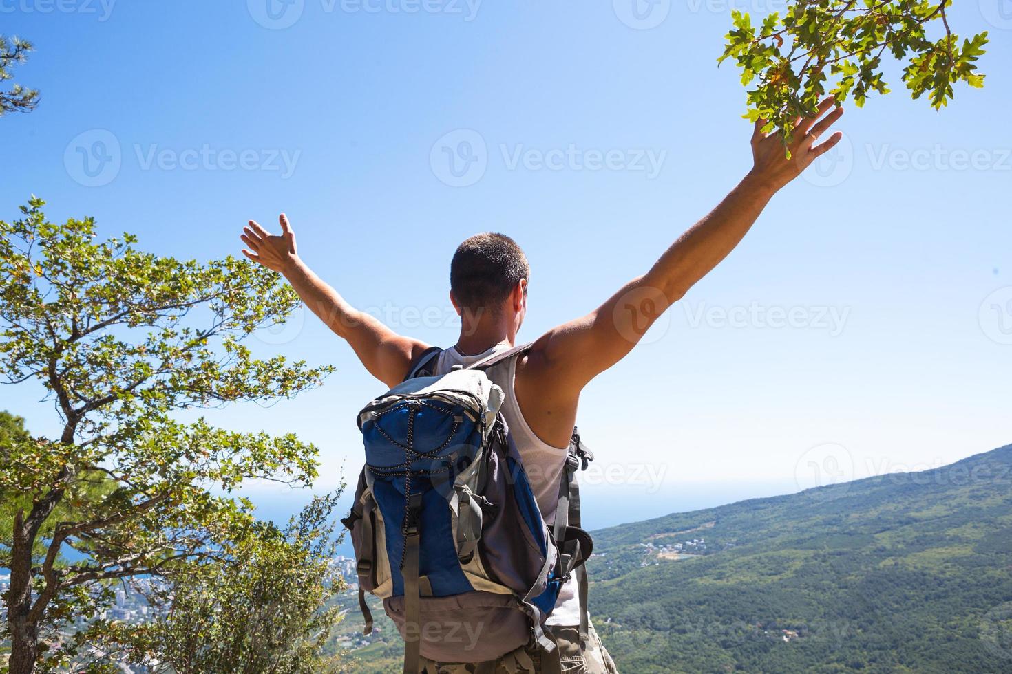 Male tourist with a backpack on top of a mountain with his hands raised looks at panorama of the coast, city, rejoices in freedom. Travel, trekking, hiking, active lifestyle, sports tourism, hiking. photo