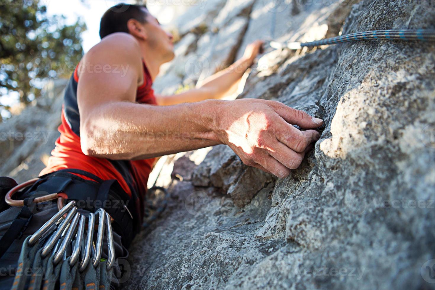 escalador en camiseta roja sube una roca gris. una mano fuerte agarró el plomo, el enfoque selectivo. fuerza y resistencia, equipo de escalada cuerda, arnés, tiza, bolsa de tiza, mosquetones, aparatos ortopédicos, cintas exprés foto