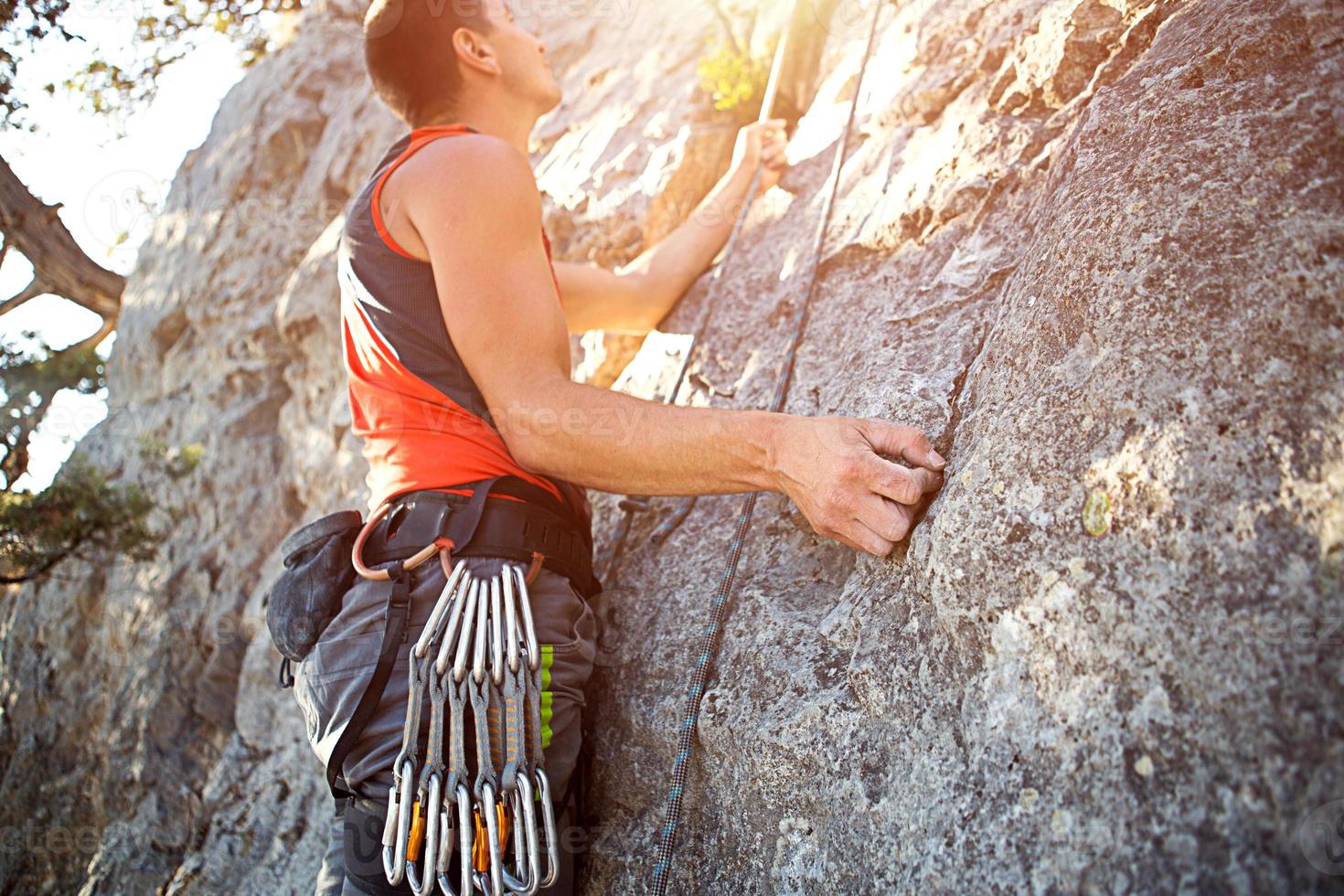 Climber in red t-shirt climbs a gray rock. A strong hand grabbed the lead, selective focus. Strength and endurance, climbing equipment rope, harness, chalk, chalk bag, carabiners, braces, quickdraws photo