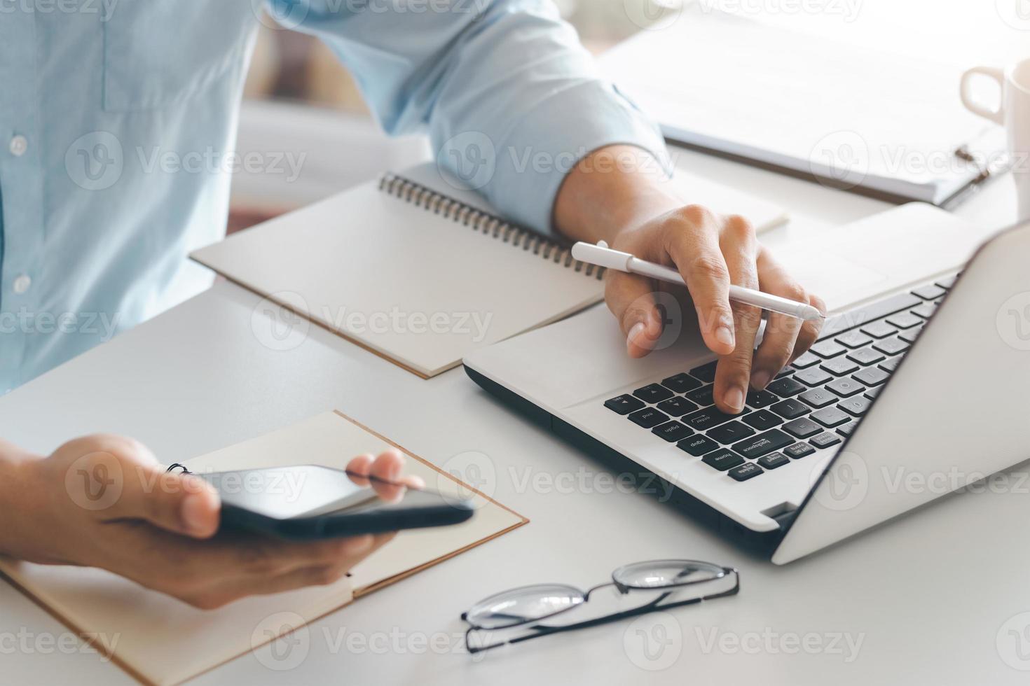 young man using laptop computer and mobile phone When looking for financial information in business, work at the desk. Writing with a pen, studying remotely from home and working from home. photo