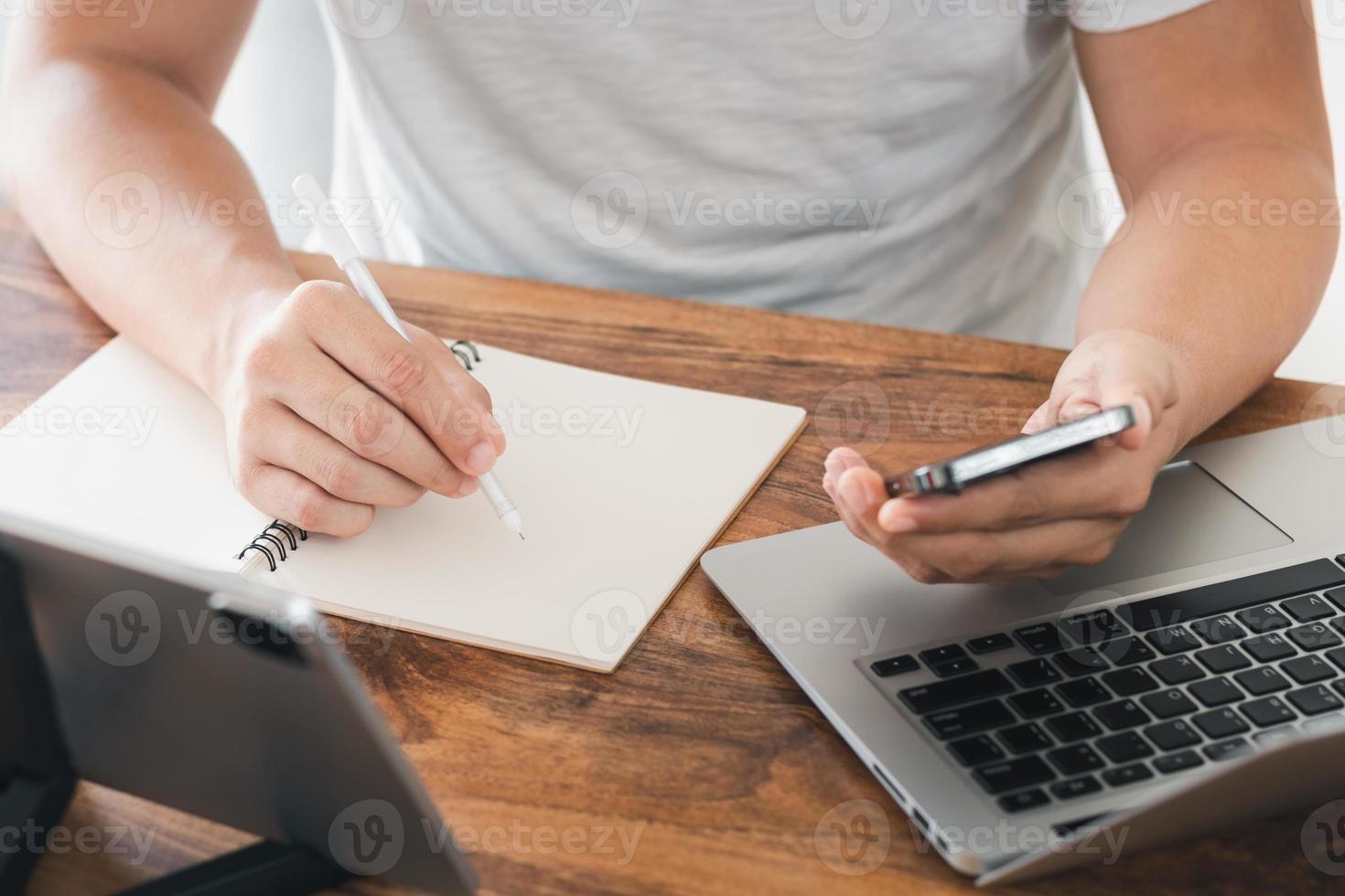 young man using laptop computer and mobile phone When looking for financial information in business, work at the desk. Writing with a pen, studying remotely from home and working from home. photo