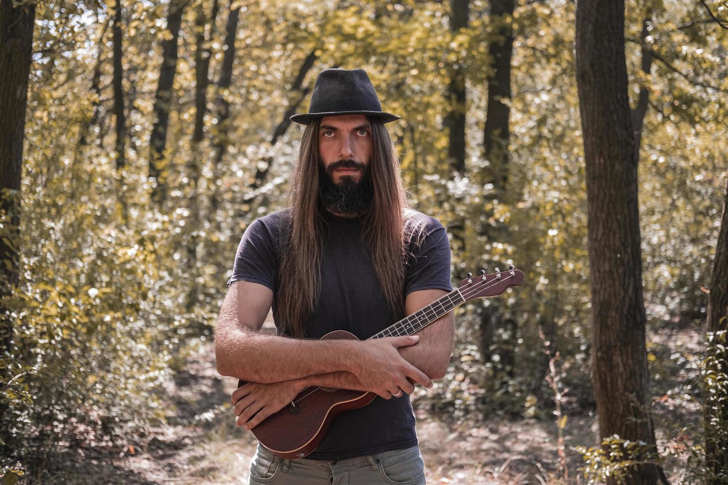 hombre barbudo con pelo largo y sombrero posando con ukelele en el bosque foto