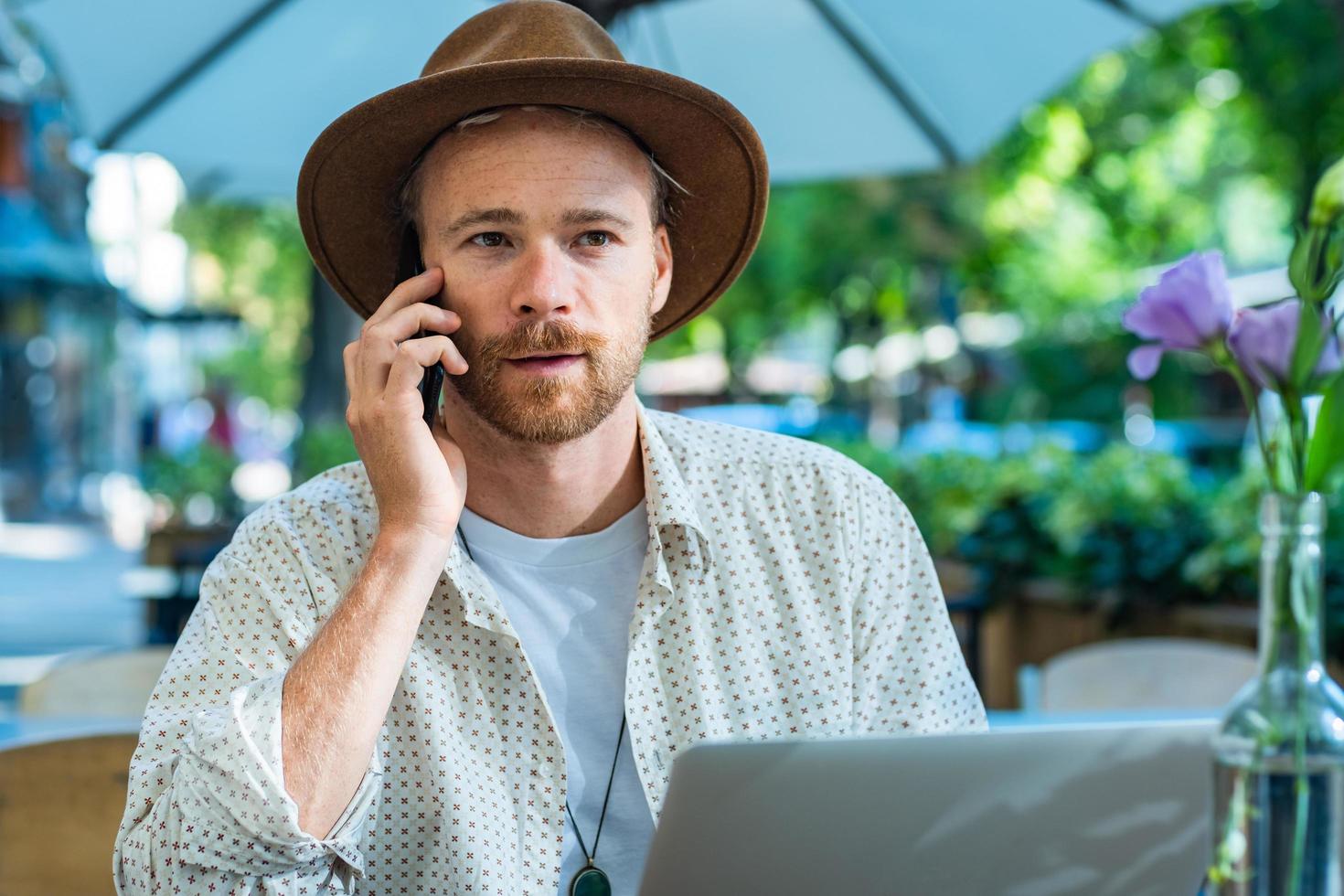 joven hipster elegante con sombrero trabaja con una laptop al aire libre en el café de la calle foto