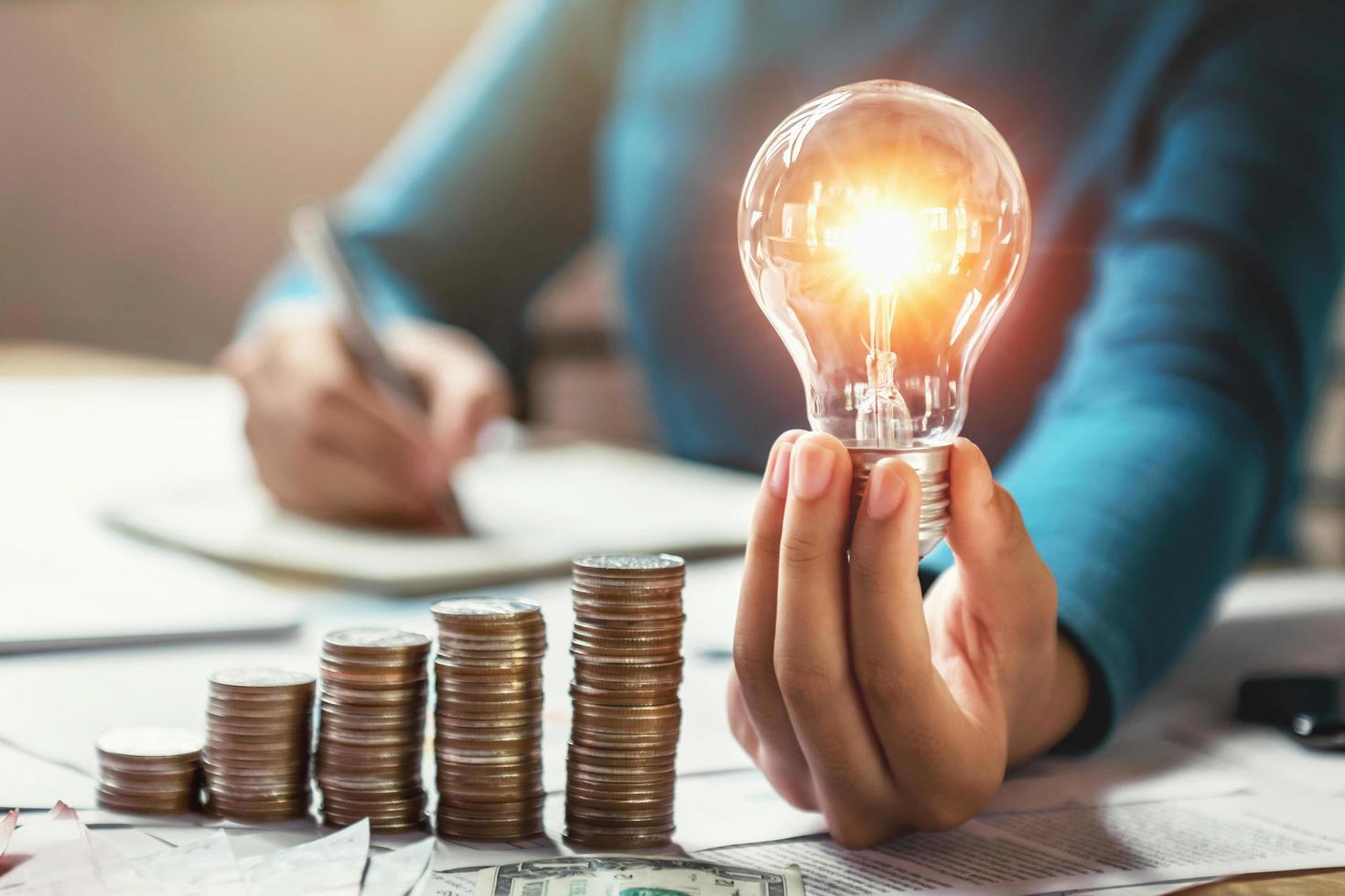 mano de mujer de negocios sosteniendo bombilla con pila de monedas en el escritorio. concepto de ahorro de energía y dinero foto