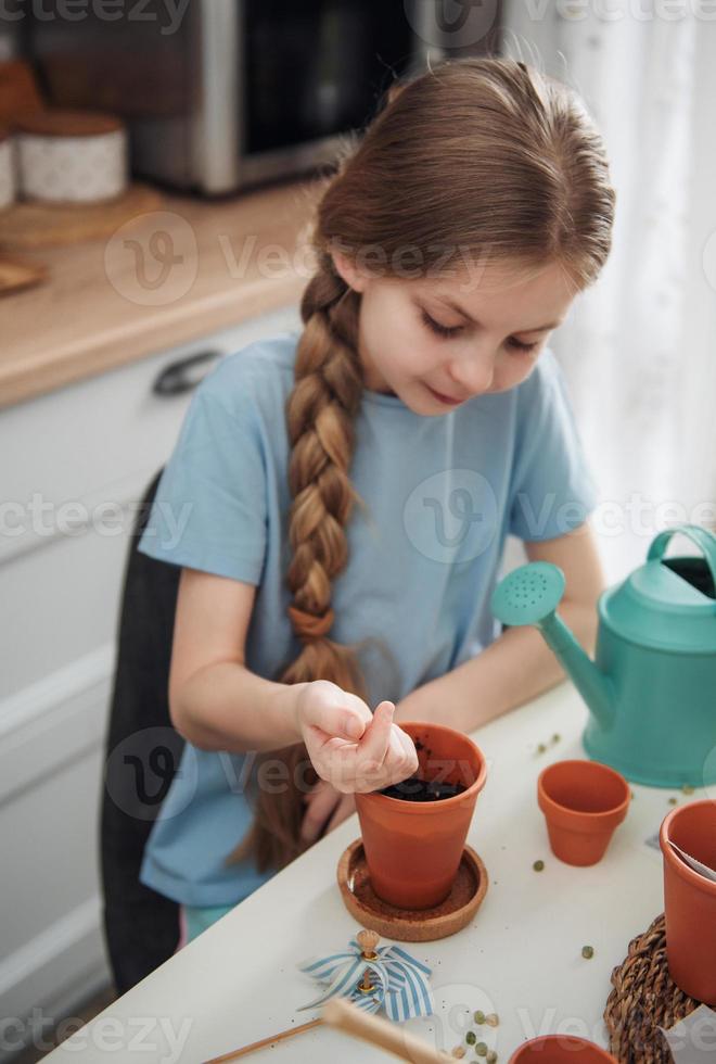 niña sentada en la mesa en casa, sembrando semillas en macetas. foto