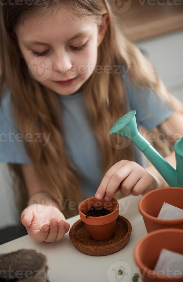 niña sentada en la mesa en casa, sembrando semillas en macetas. foto