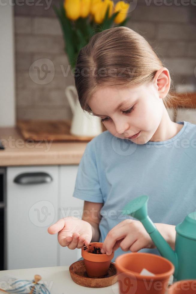 niña sentada en la mesa en casa, sembrando semillas en macetas. foto