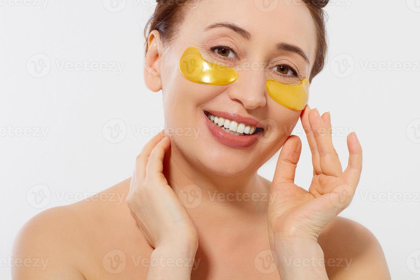 Macro female face. Beauty portrait of middle age woman with wrinkles and a gold patch under eye isolated on white background. Collagen mask and spa concept. Copy space. Summer skin care photo