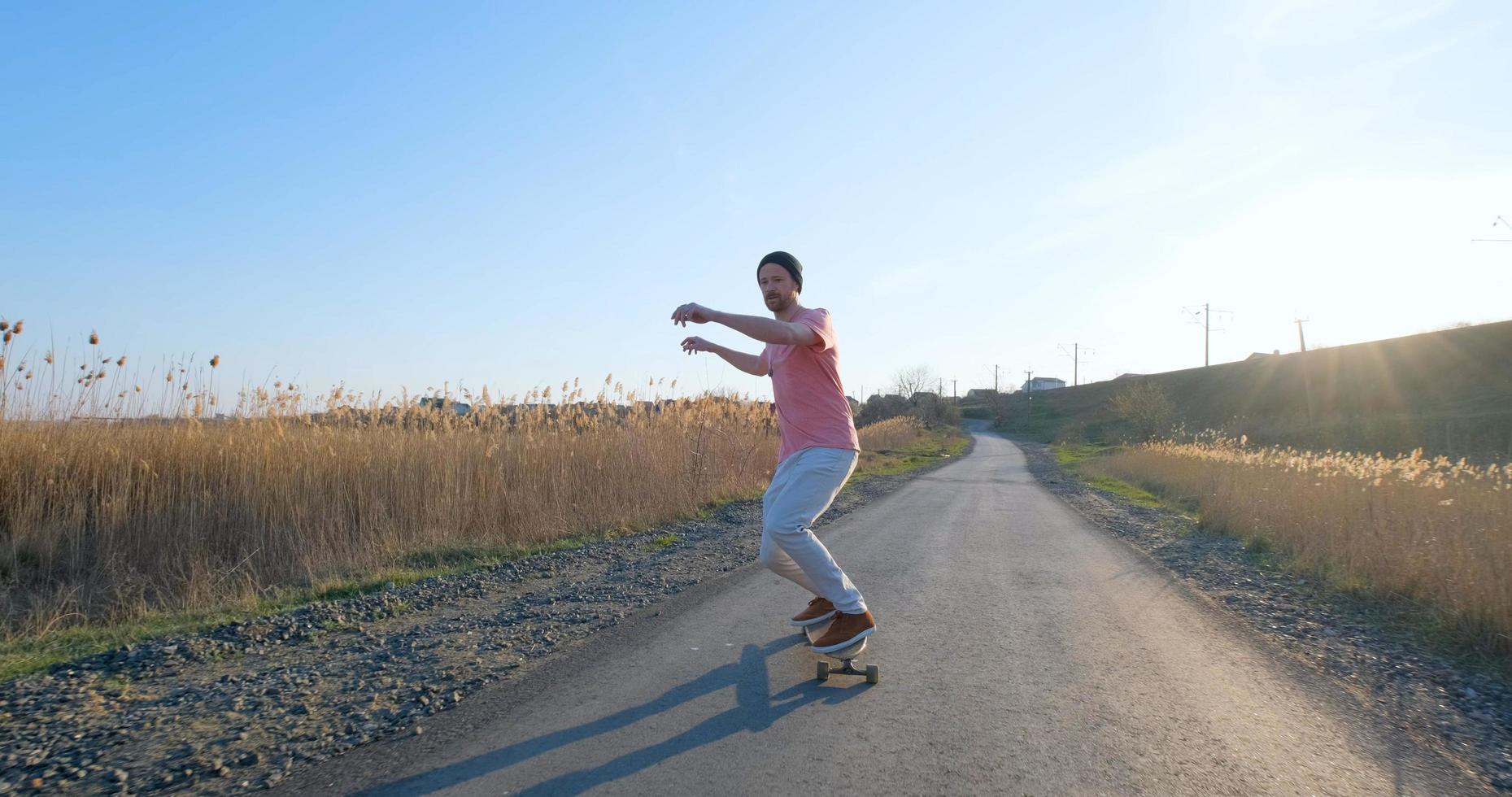 paseo masculino joven en patineta longboard en la carretera del país en un día soleado foto