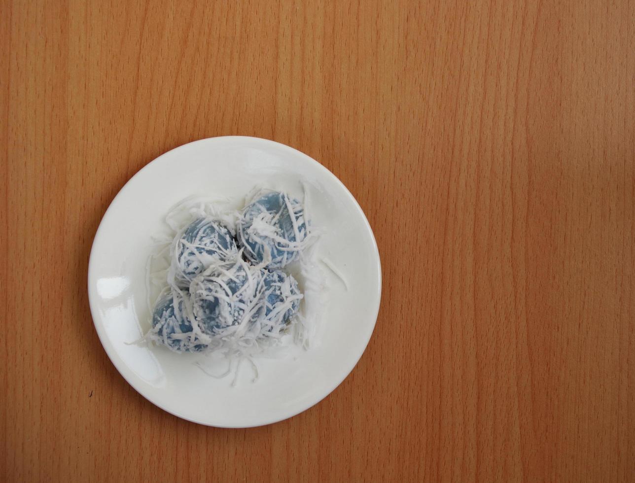 Glutinous rice ball with palm sugar roll up grated ripe coconut on white round dish and light brown wood broad background. Thai traditional dessert. photo