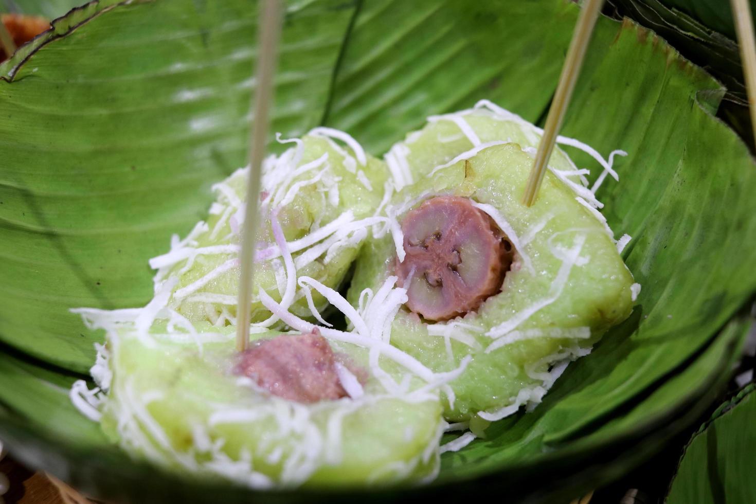trozos de plátano envueltos en arroz pegajoso están en un plato redondo de hojas de plátano, servidos con azúcar y coco rallado, tailandia. foto