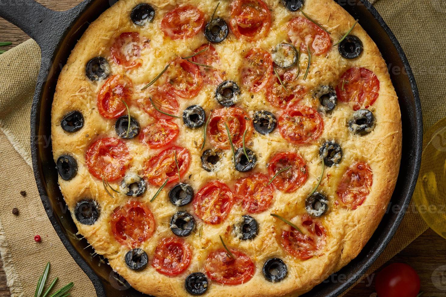 Focaccia, pizza in skillet, italian flat bread with tomatoes, olives and rosemary. Wooden table photo