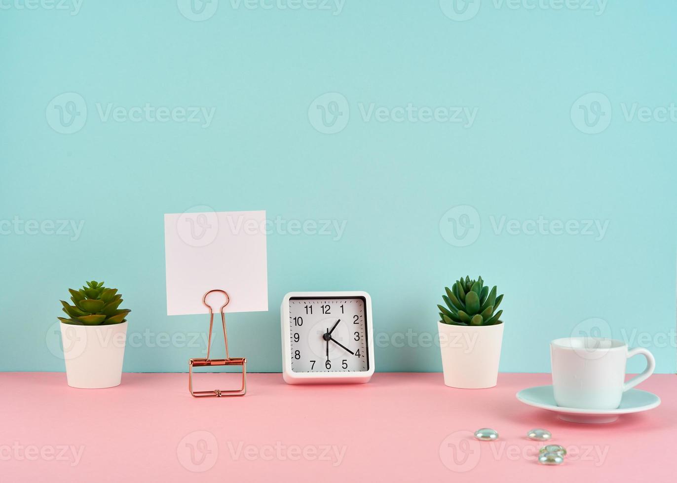 Mockup with white frame, note, alarm, cup of coffee or tea on pink table against blue wall photo