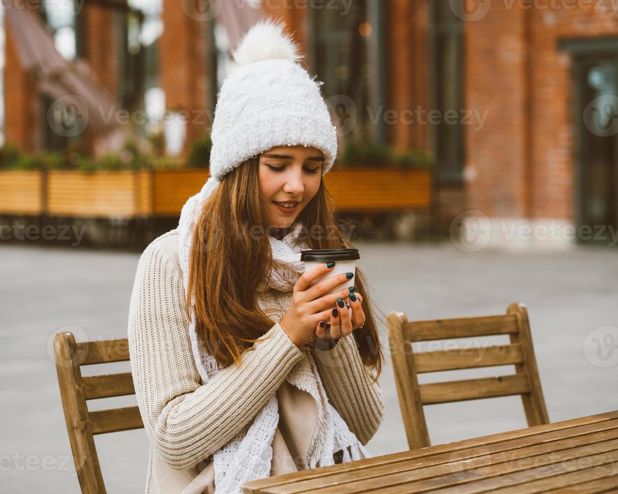 Beautiful young girl drinking coffee, tea from plastic mug in autumn, winter. A woman with long hair in warm clothes sitting in street cafe, warmed by hot drink photo