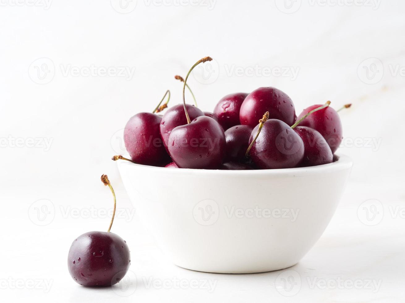 red dark sweet cherries in white bowl on stone white table, side view. photo