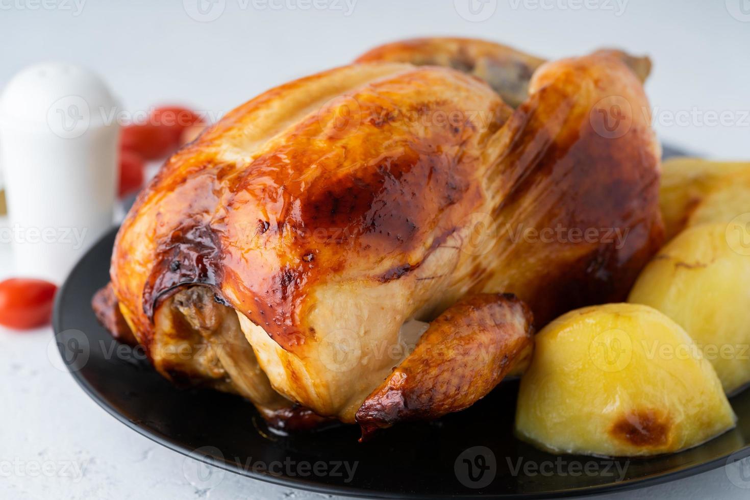 Grilled whole chicken in black plate on white table, baked meat with potatoes. Side view, close up photo
