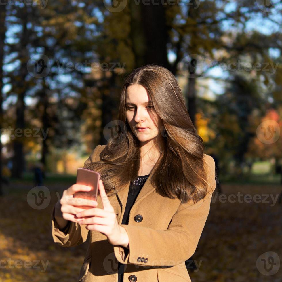 Beautiful teen girl in the Park, holding smartphone and chatting online on Internet, takes a selfie. Young woman with long hair photo