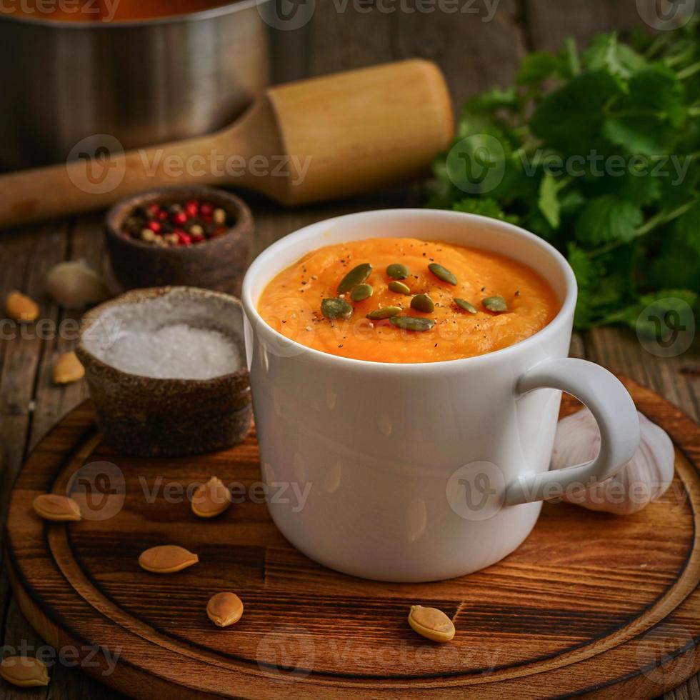 sopa de crema de calabaza en una taza sobre una mesa de madera marrón, vista lateral. puré vegetariano dietético en tabla de cortar con perejil, ajo. foto