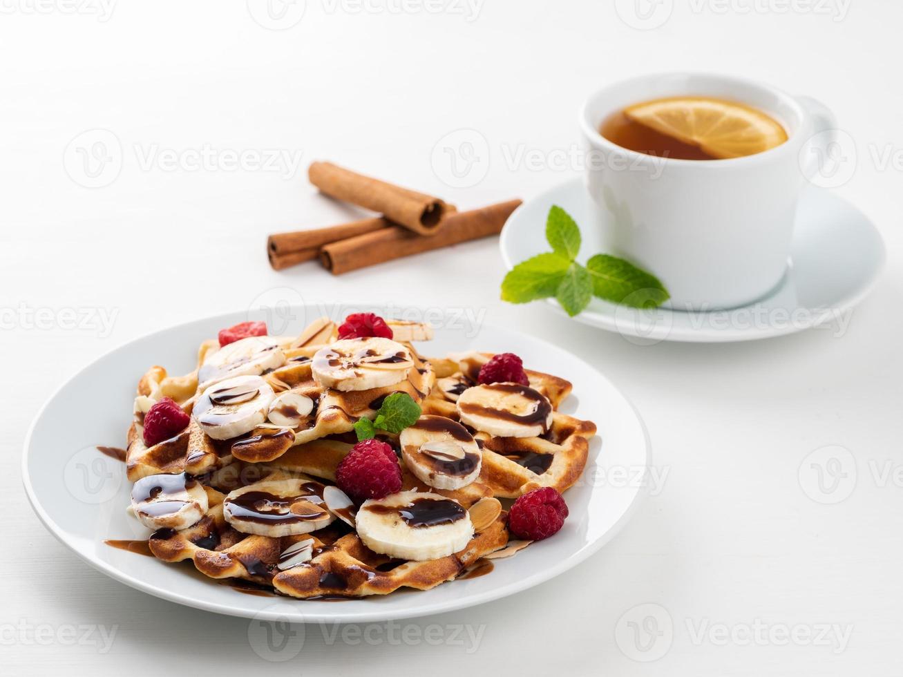 Belgian waffles with raspberries, banana, chocolate syrup. Breakfast with tea on white background, side view photo