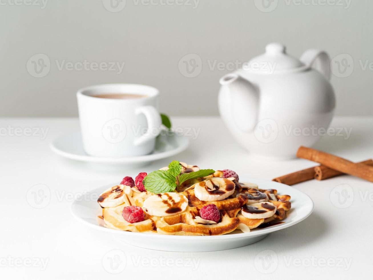 Belgian curd waffles with raspberries, banana, chocolate syrup. Breakfast with tea on white background, side view photo