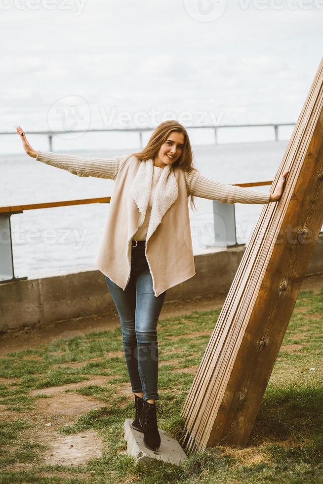 Happy young girl standing on waterfront at pier in port, enjoying life and waving his arms. Woman with long hair smiles and enjoys moment photo