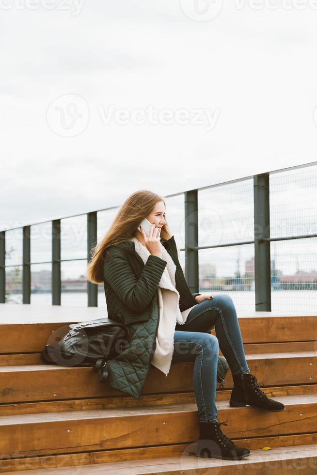 hermosa mujer con cabello largo hablando por teléfono. otoño o invierno, chica con chaqueta al aire libre sentada en escalones de madera foto