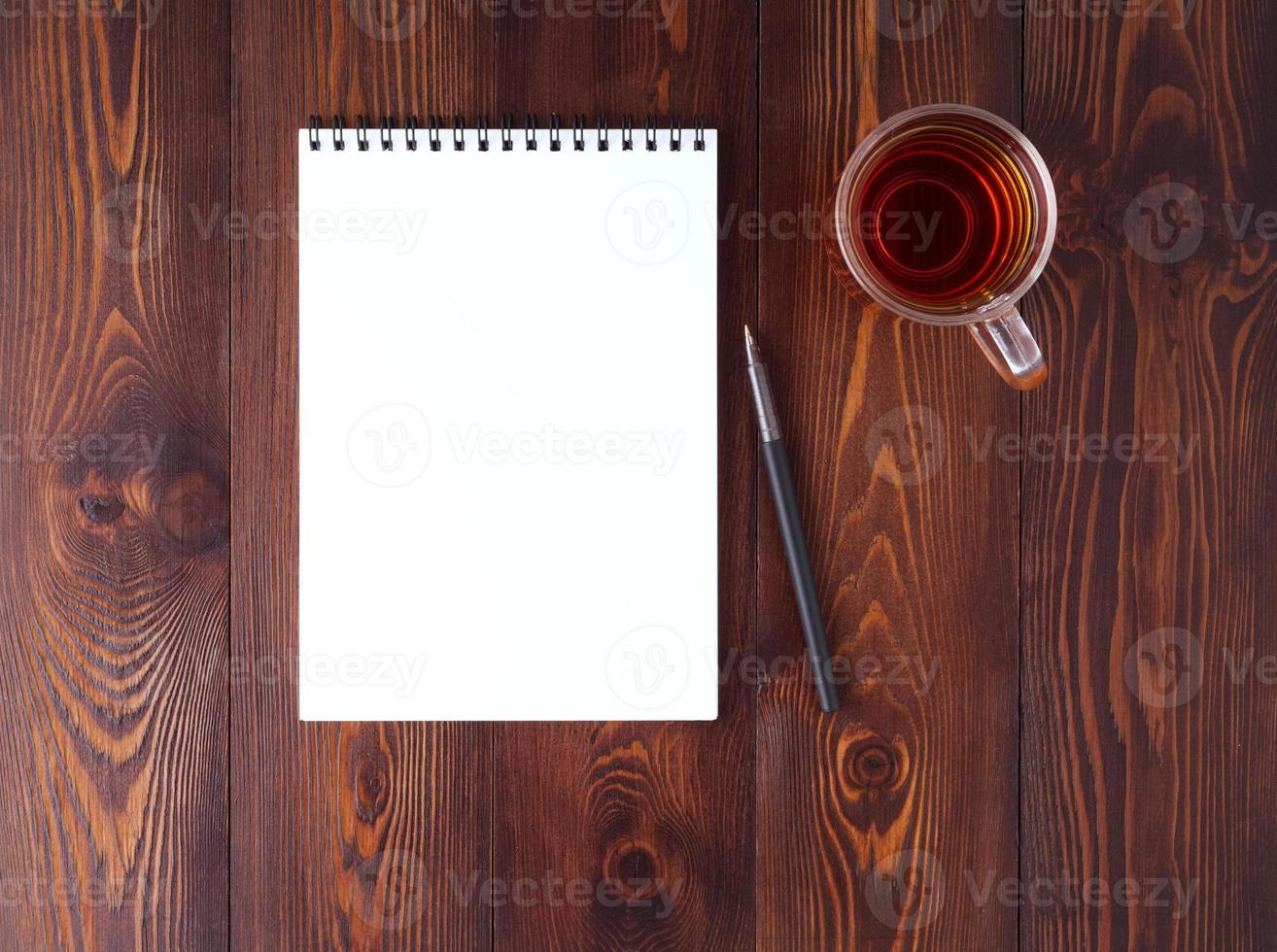The open notepad with spiral , pencil, cup of tea on dark brown wooden table, top view photo
