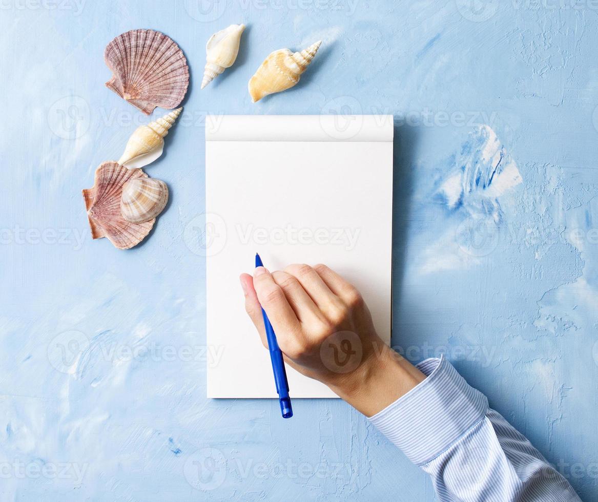woman writes in notebook on stone blue table, Mock up with frame of seashell, top view, planning holiday by sea photo