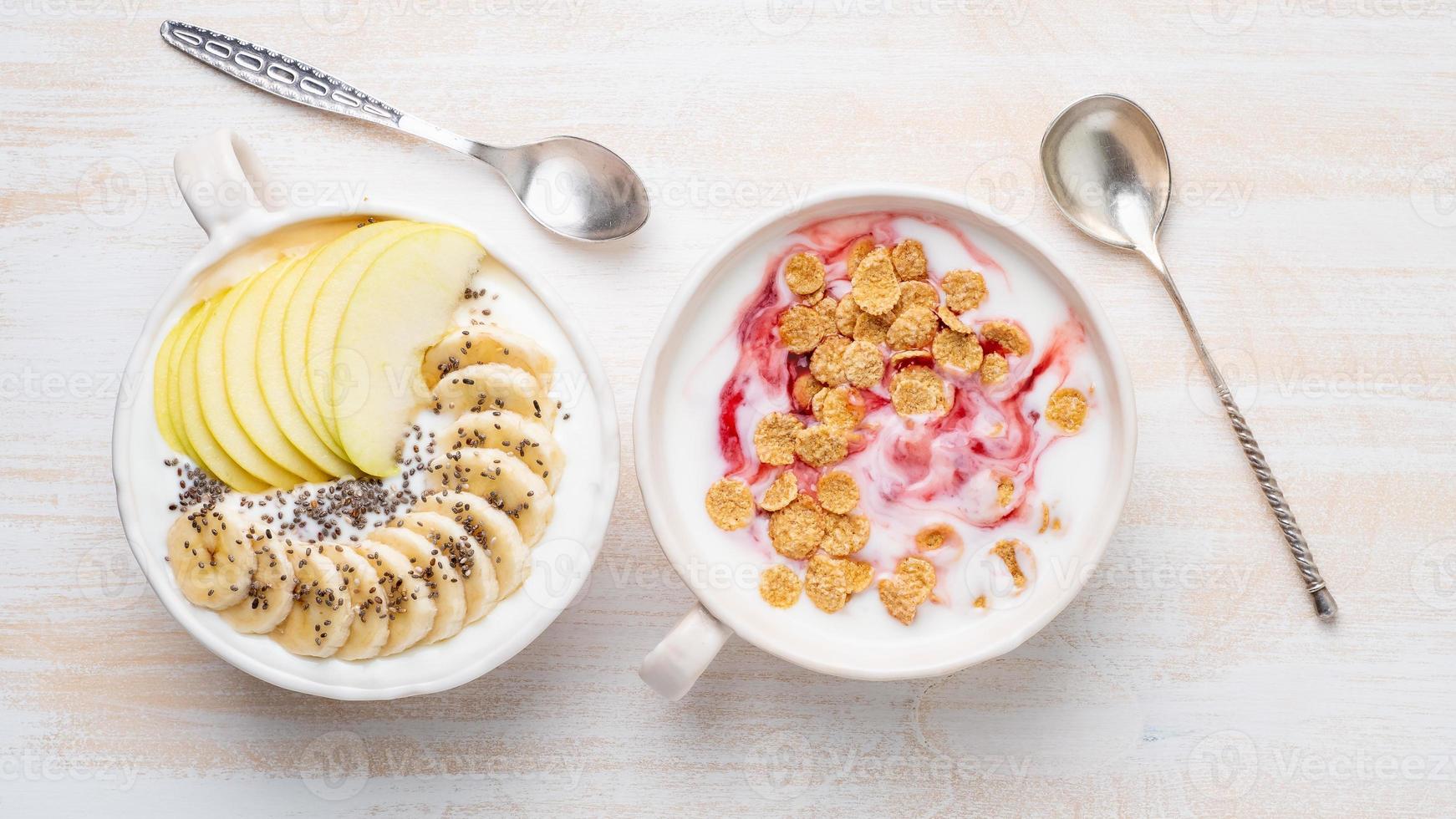 Two greek yogurt with jam, apple, muesli, chia seeds and banana in white bowl on white wooden table, top view, banner photo