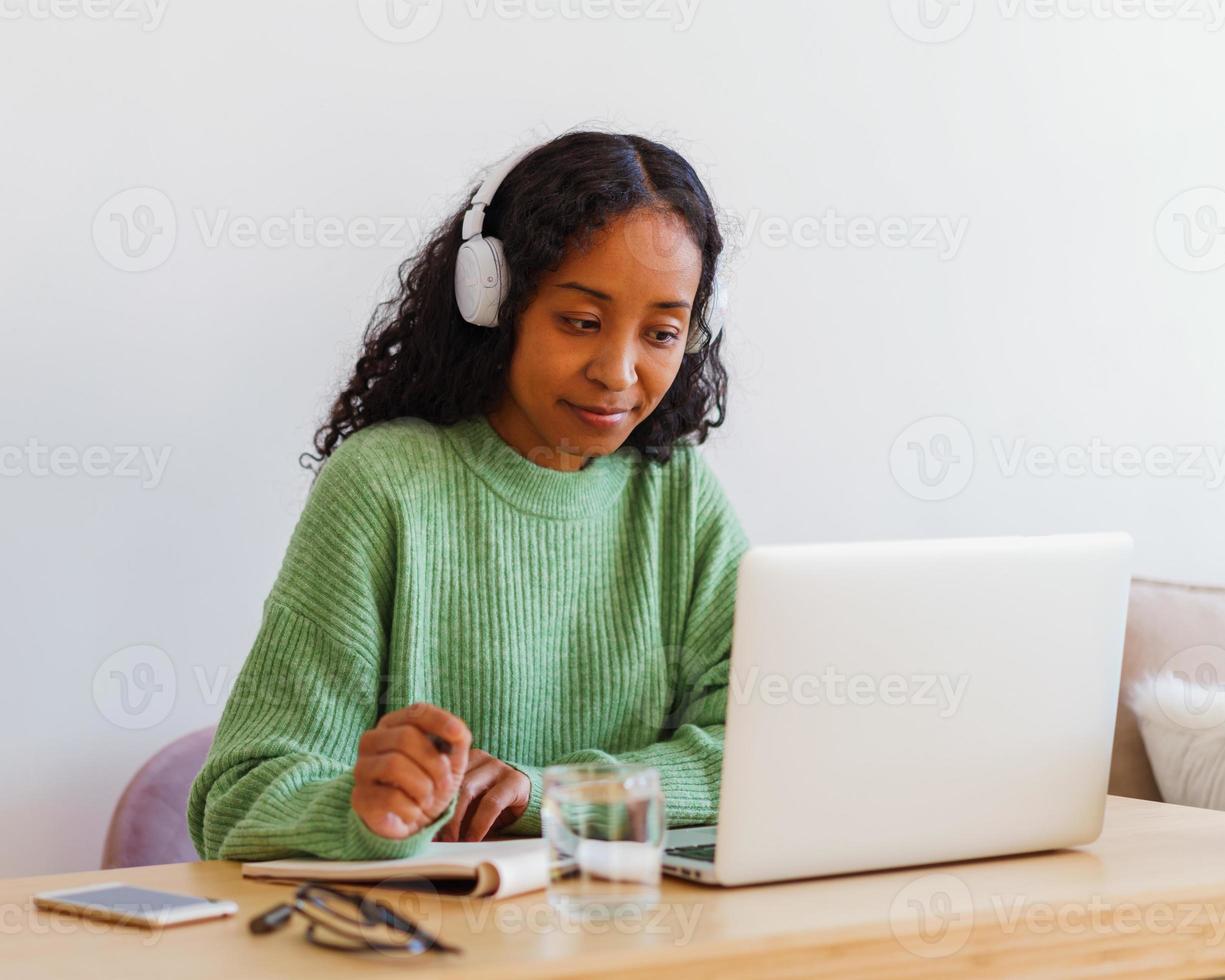 joven afroamericana trabajando en una laptop con auriculares mientras toma notas en un cuaderno foto