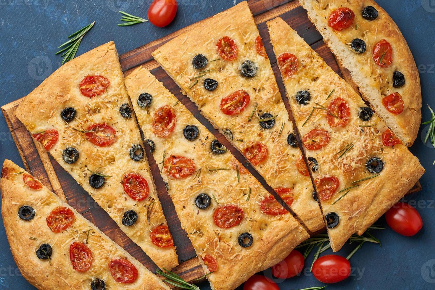 Focaccia, pizza, italian flat bread with tomatoes, olives and rosemary on dark blue table, top view photo