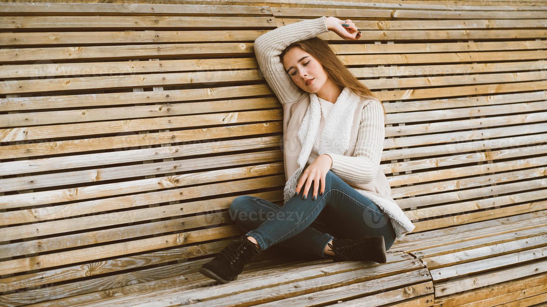 Beautiful young girl with long brown hair sits on wooden bench made of planks and rests, relaxes and reflects. Outdoor photo shoot with attractive woman in winter or autumn
