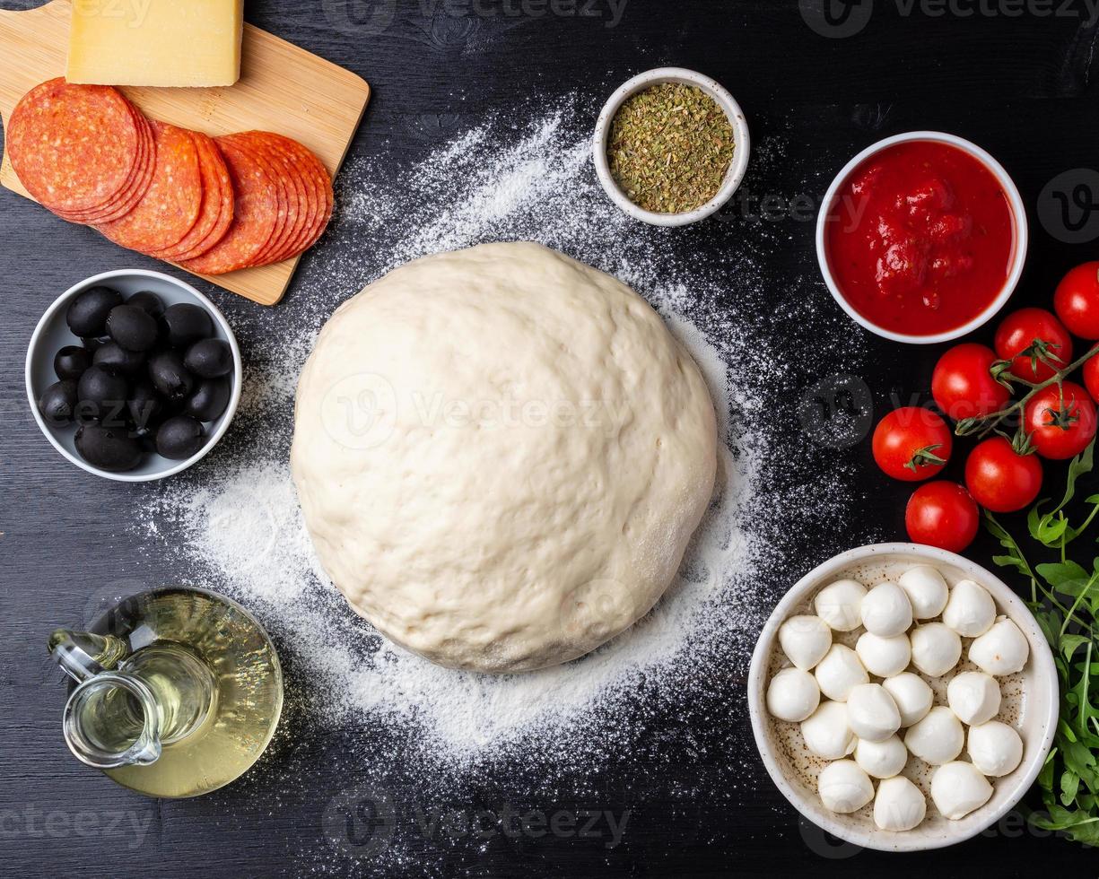 masa cruda para pizza con ingredientes y especias sobre fondo negro oscuro, vista superior, puesta plana foto