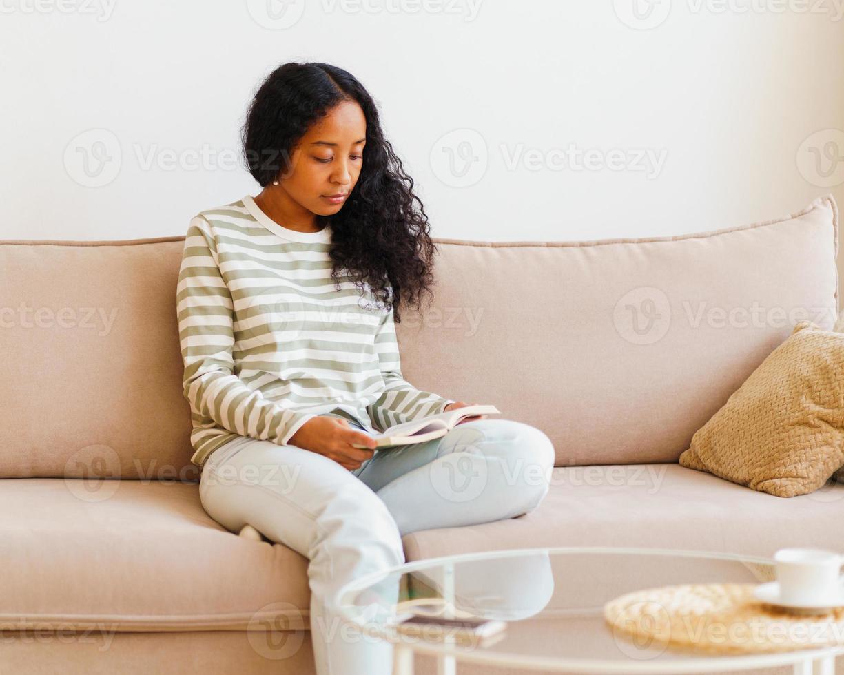 Beautiful African-American female sitting on couch and reading book. Spending time at home alone photo