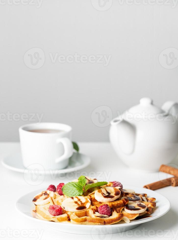 Belgian curd waffles with raspberries, banana, chocolate syrup. Breakfast with tea on white background, side view, vertical photo