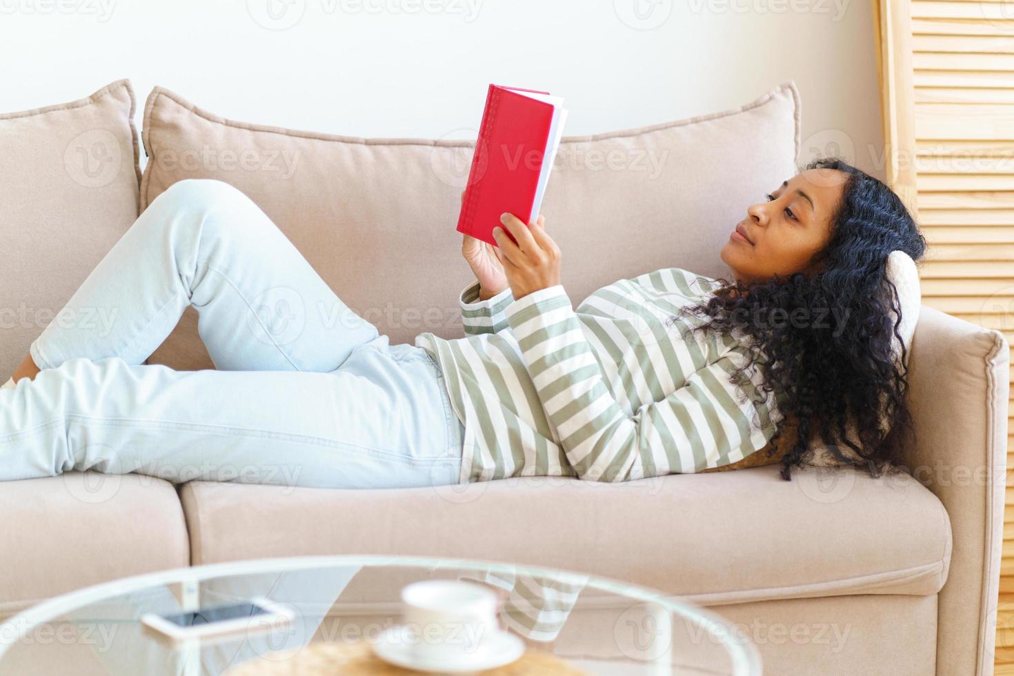 afroamericano acostado en el sofá y leyendo un libro con tapa roja. estilo de vida lento foto