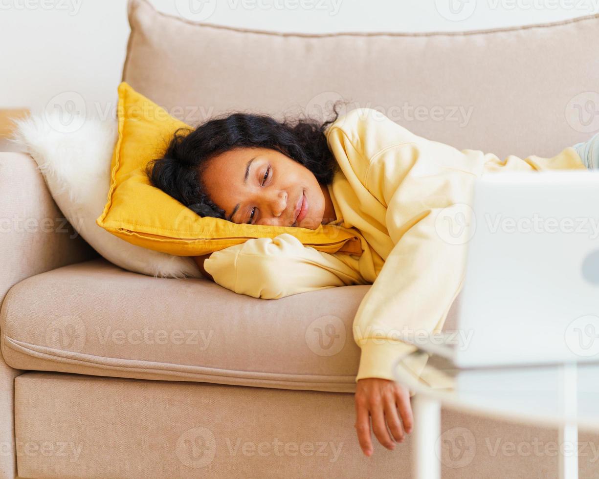 mujer afroamericana feliz y sonriente acostada en el sofá y viendo películas en la computadora portátil foto