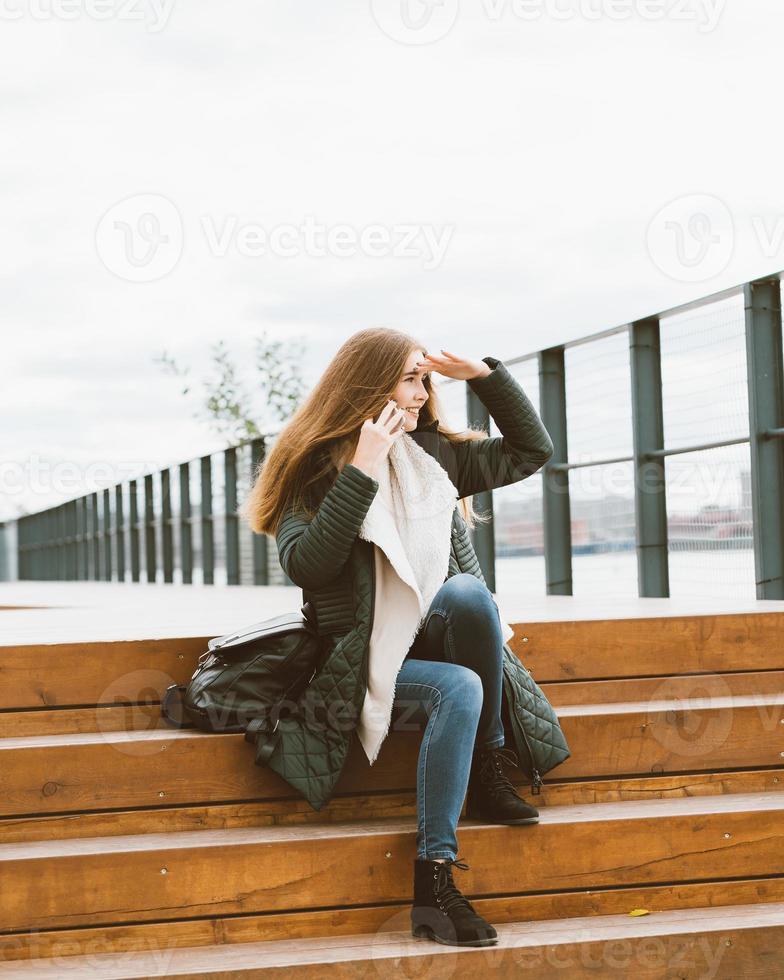 Beautiful woman with long hair talking on phone and looking for something in distance. Autumn or winter, girl in outdoor jacket sitting on wooden steps photo