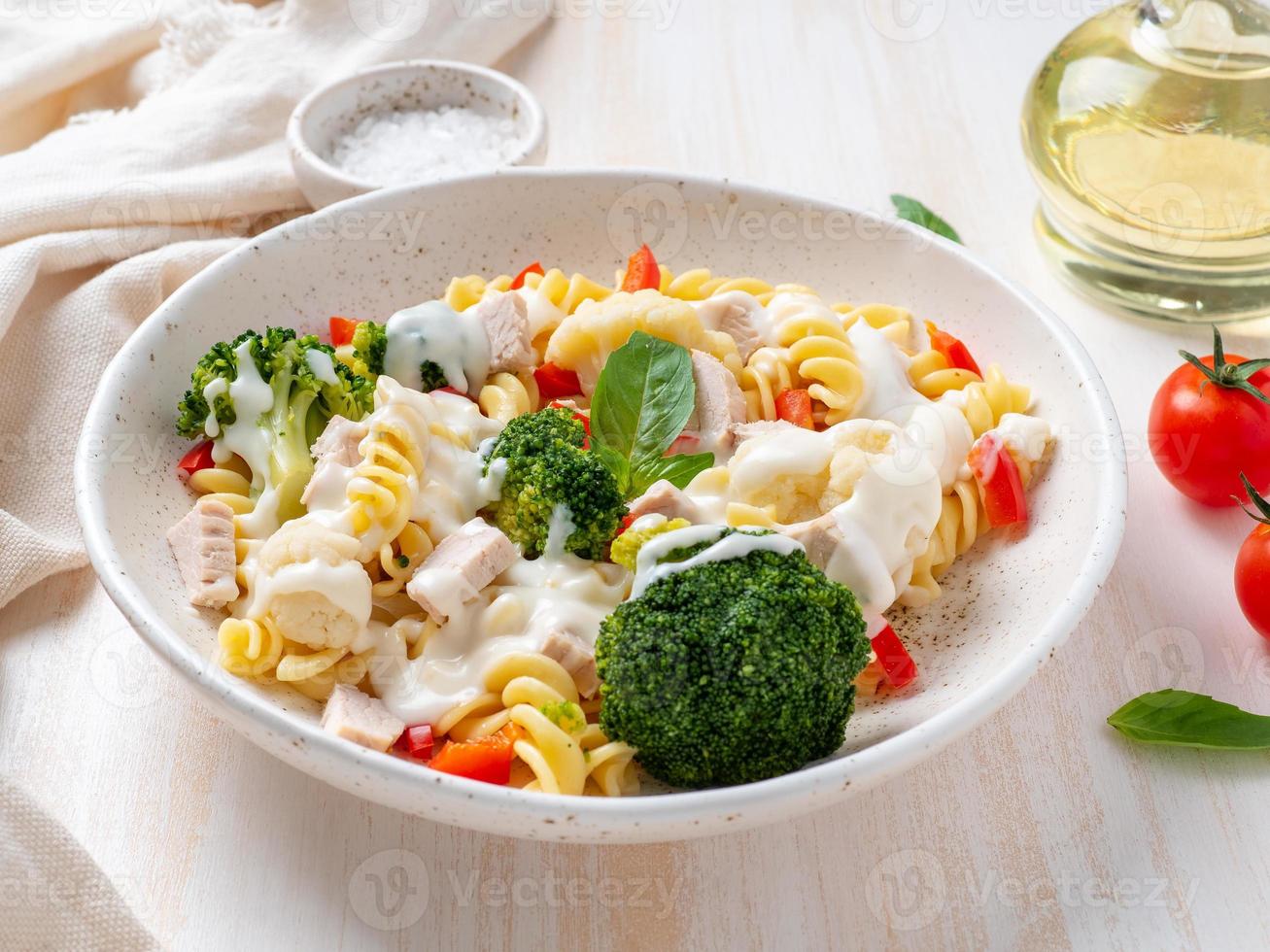 Pasta fusilli with vegetables, boiled steamed meat, white sauce on white wooden table, low-calorie food, low-fat diet, side view photo